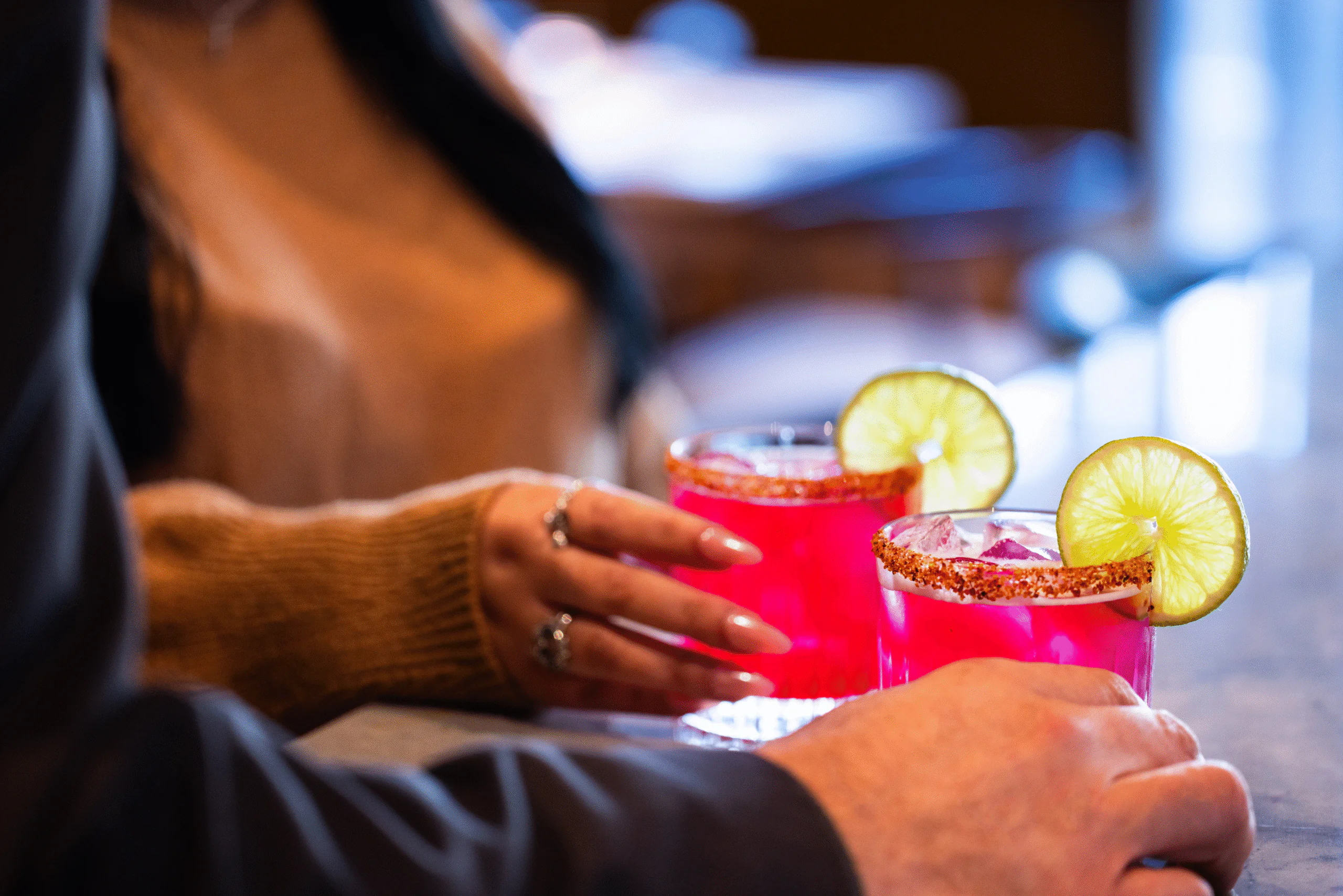 Two people holding bright pink cocktails with chili rims and lime slices, sitting at a bar with a blurred background.