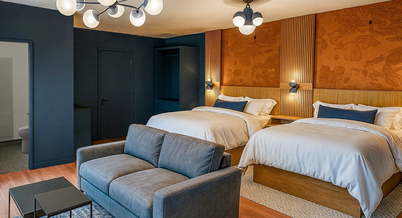 Modern hotel room with two double beds, a gray sofa, wooden floors, dark blue walls, and contemporary lighting fixtures.