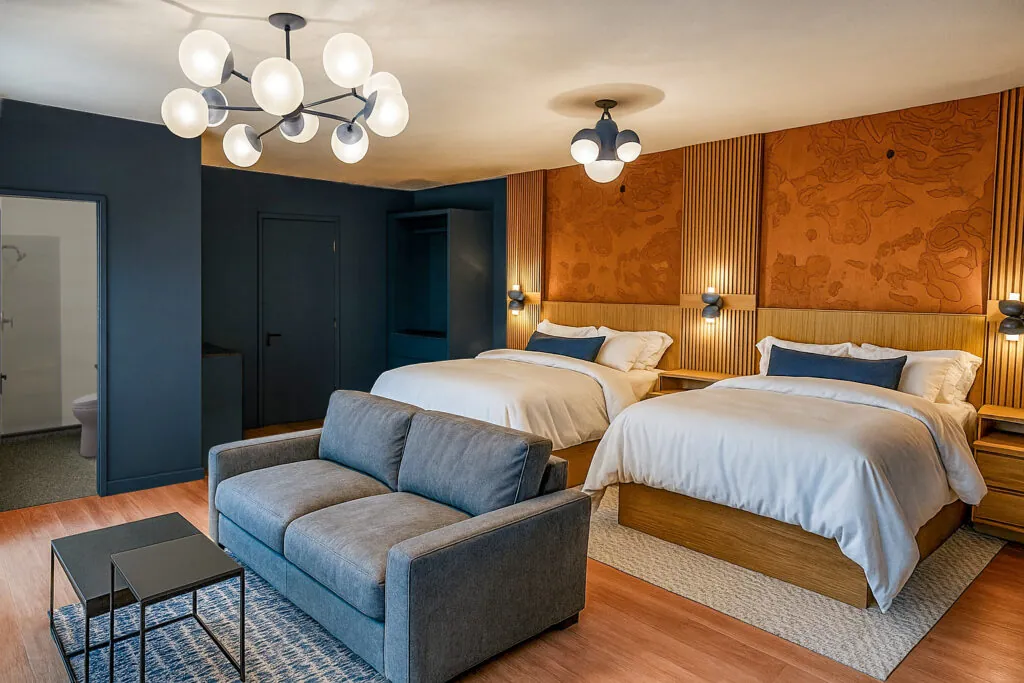 Modern hotel room with two queen beds, a gray sofa, side tables, warm lighting, and contemporary decor in shades of blue, brown, and white.