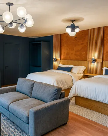 Modern hotel room with two queen beds, a gray sofa, side tables, warm lighting, and contemporary decor in shades of blue, brown, and white.