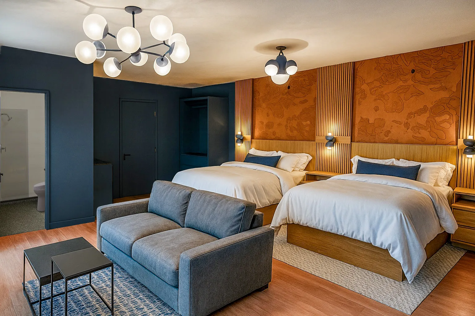 Modern hotel room with two queen beds, a gray sofa, side tables, warm lighting, and contemporary decor in shades of blue, brown, and white.
