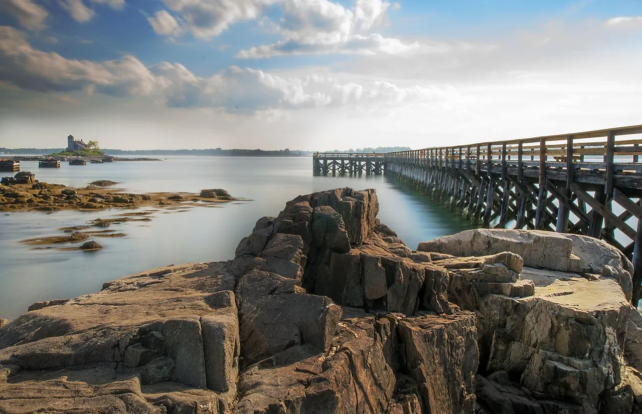 Rocky shoreline with a wooden pier extending over calm water under a partly cloudy sky, with an island visible in the distance.