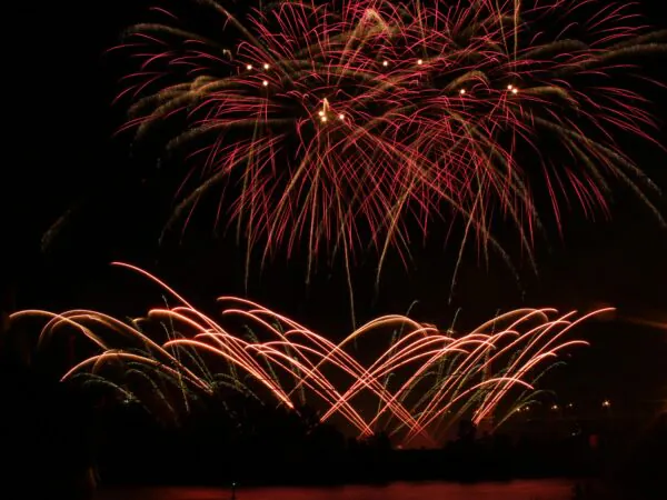 Red and gold fireworks burst and arc over a dark night sky, with faint silhouettes of trees and buildings below.