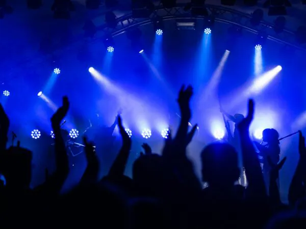 Silhouetted crowd claps and cheers at a concert, with bright blue stage lights illuminating the band performing in the background.