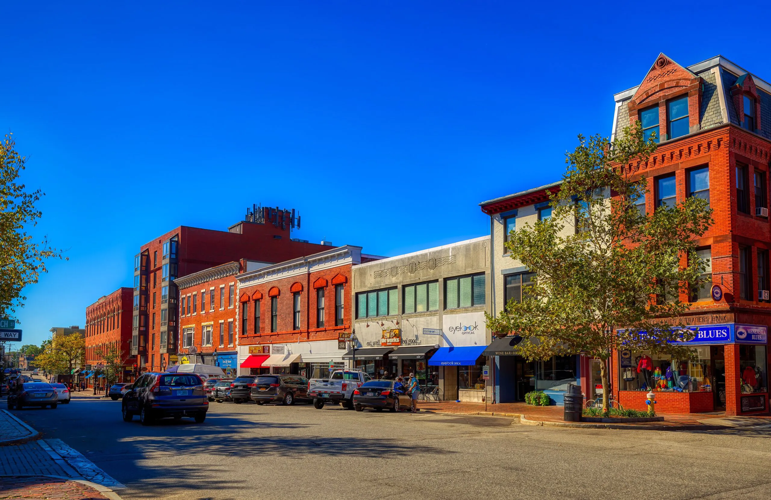 A vibrant small town street with brick buildings, parked cars, storefronts, and a clear blue sky on a sunny day.