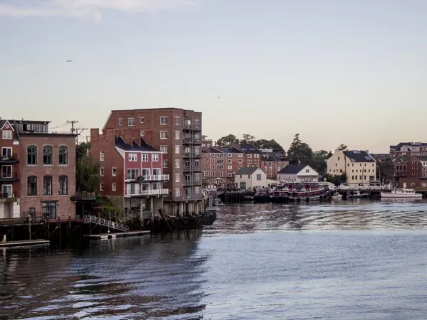 Brick buildings line a waterfront at dusk, with reflections in the calm water and boats docked along the shore under a clear sky.