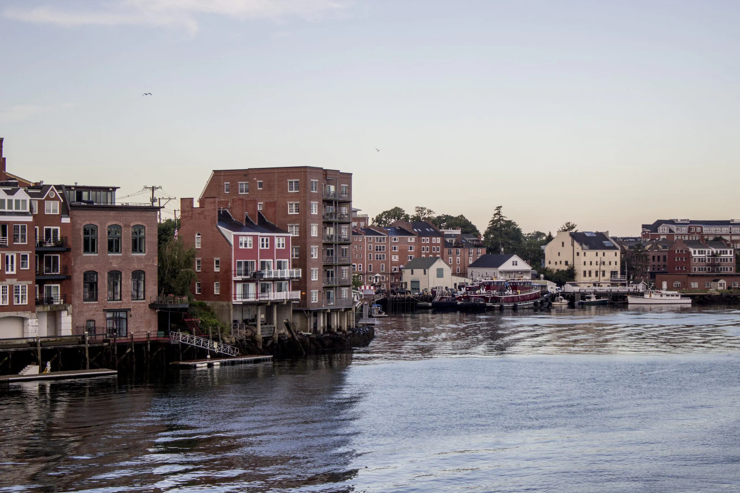 Brick buildings line a waterfront at dusk, with reflections in the calm water and boats docked along the shore under a clear sky.
