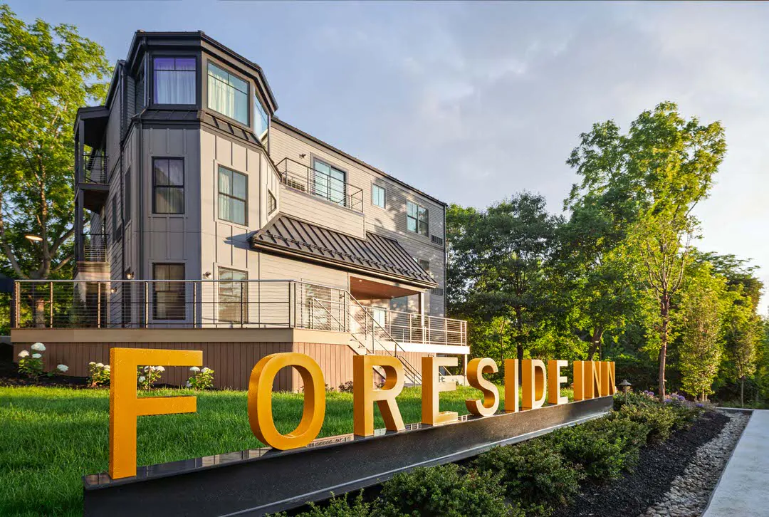 The exterior of the Foreside Inn features a multi-story, modern grey building with balconies, set behind a prominent, large gold sign reading 'FORESIDE INN' on a grassy lawn during late afternoon, sunlit conditions.