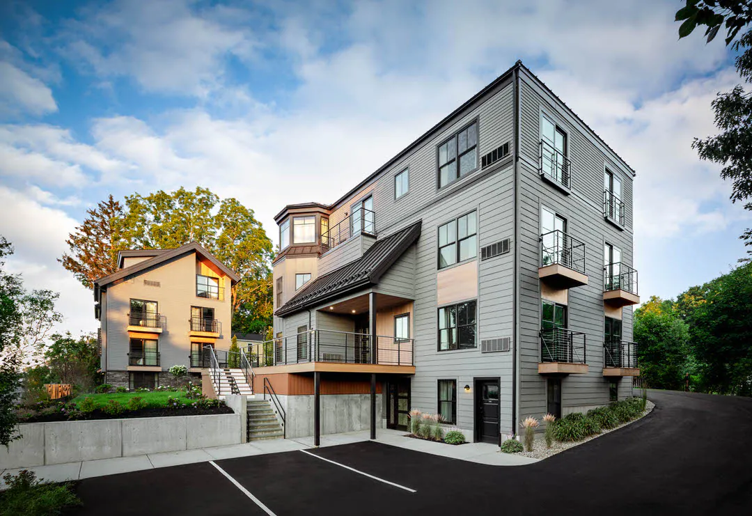 A modern apartment building with gray siding, multiple balconies, and a paved parking lot in the foreground. A smaller, matching residential building stands in the background beneath a blue sky with soft white clouds.