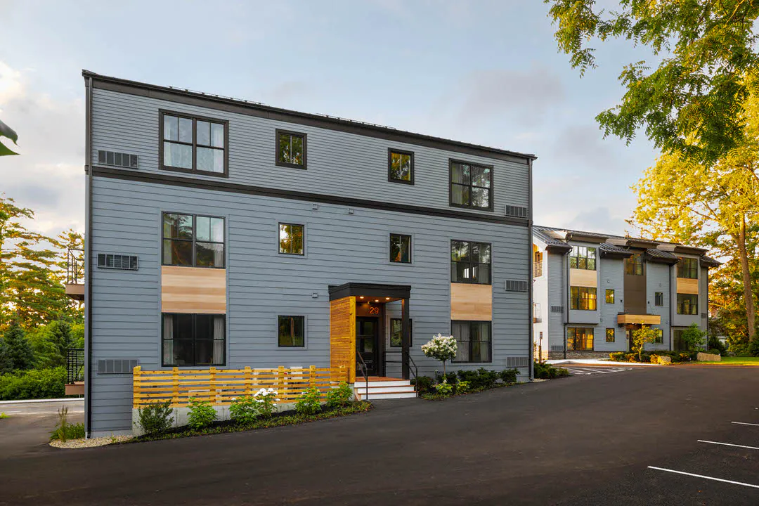 Modern residential building with blue-grey exterior siding, light wood paneling, and a black entryway marked with the number 29. The scene includes a paved driveway and lush green trees under a soft, golden-hour sky.