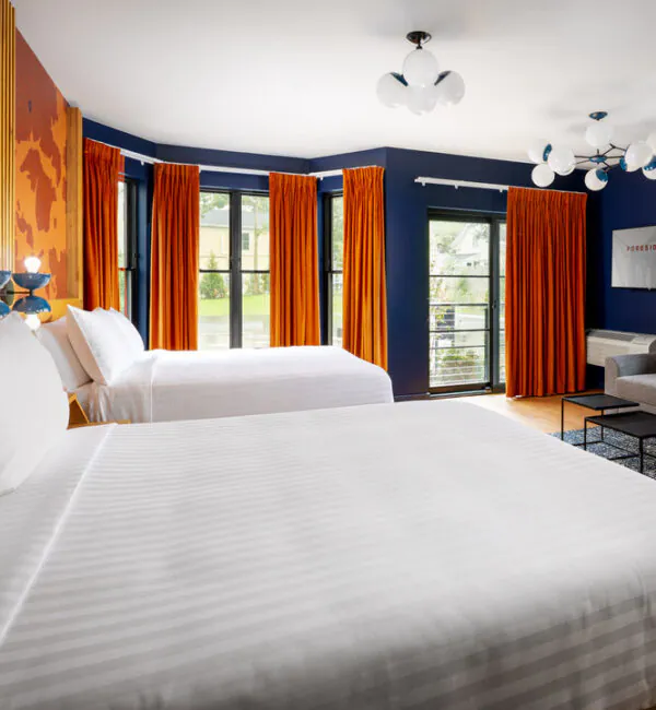A stylish hotel room featuring two beds with white striped linens, deep blue walls, and bright orange floor-to-ceiling curtains. There is a grey sofa and black coffee tables in the corner, modern globe lights, and a TV on the wall.