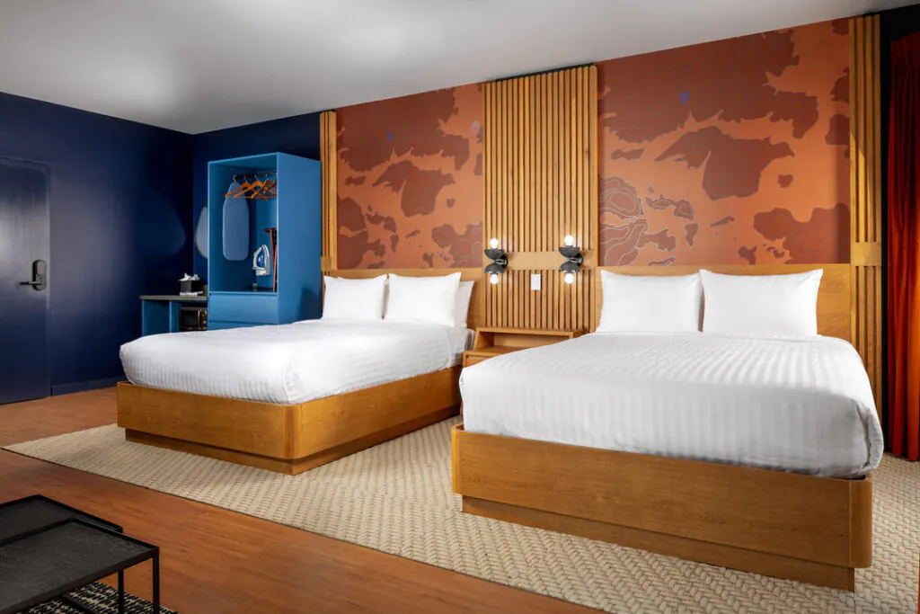 A modern hotel room featuring two beds with white linens against a backdrop of a wooden slat headboard and an orange wall with a map-like graphic. To the left is a navy blue accent wall and a blue open closet containing an iron and board.