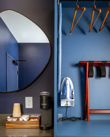 A hotel room corner features a dark blue wall with an irregularly shaped mirror. A coffee station includes a Keurig machine, supplies, and a tissue box. To the right, a closet area contains clothes hangers, an iron, and a folding luggage rack.