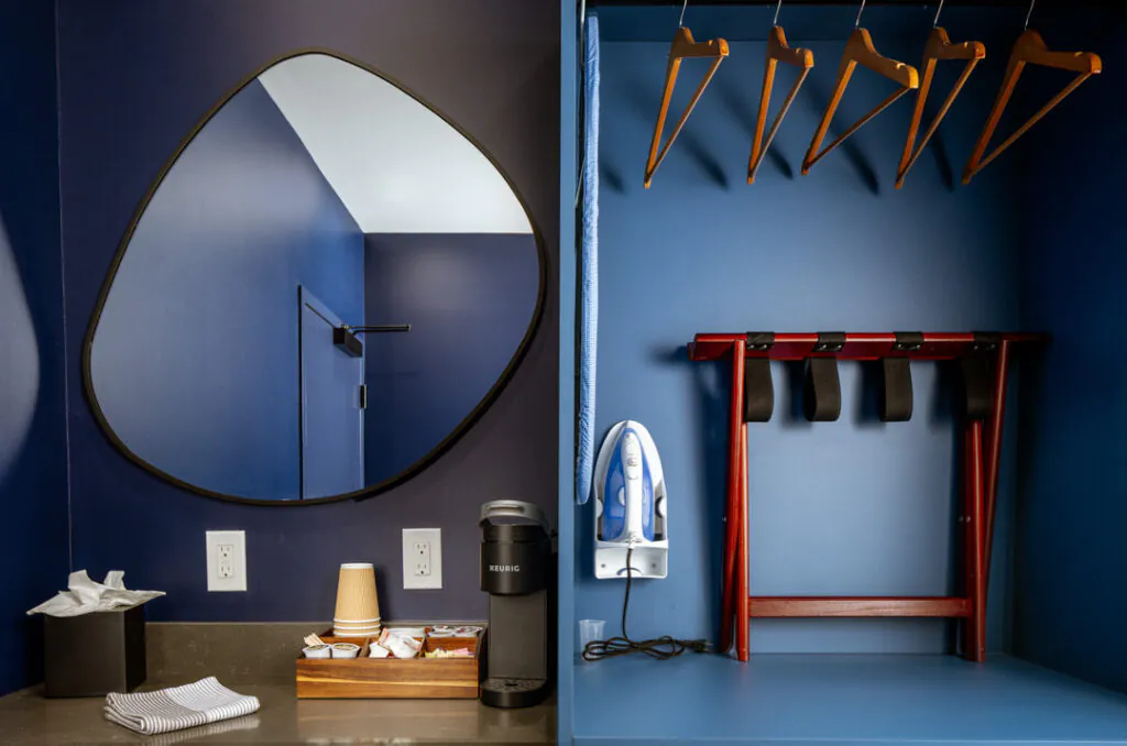 A hotel room corner features a dark blue wall with an irregularly shaped mirror. A coffee station includes a Keurig machine, supplies, and a tissue box. To the right, a closet area contains clothes hangers, an iron, and a folding luggage rack.