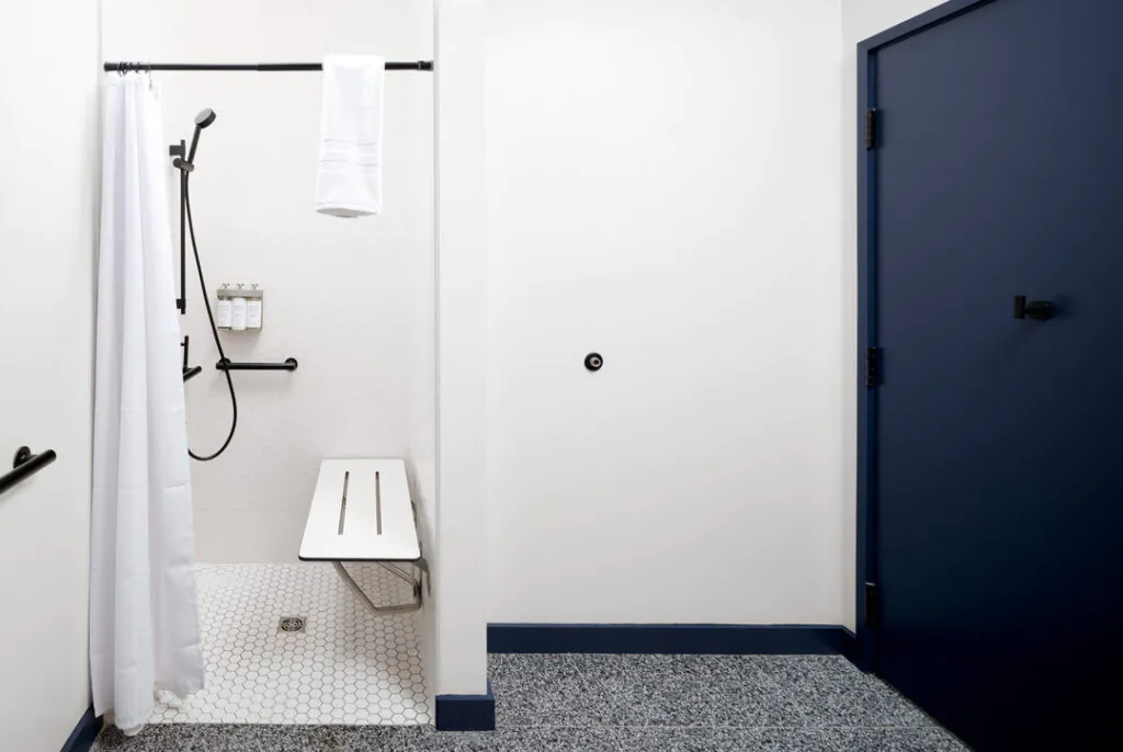 An accessible shower with a white curtain, wall-mounted seat, grab bars, handheld showerhead, and bottle dispenser. The shower area has white hexagonal tiles and is next to a solid dark blue door on a white wall with speckled grey flooring.