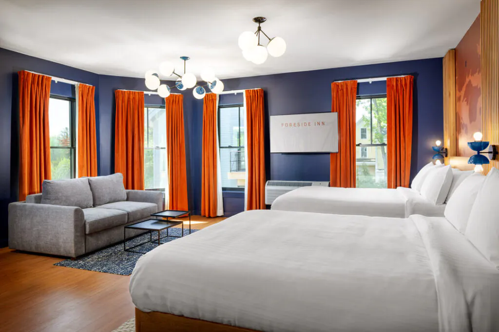 A spacious, modern hotel room featuring two queen beds with white linens, a grey sofa with black coffee tables on a patterned rug, and dark blue walls accented by vibrant orange curtains. A TV displays the Foreside Inn logo.