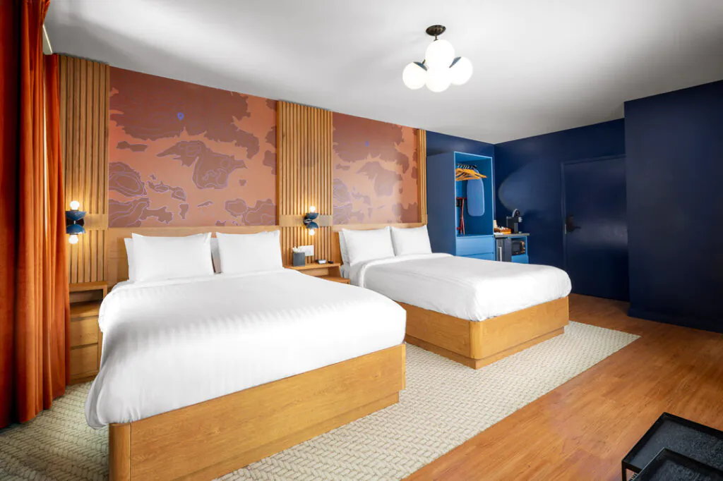 A contemporary hotel room features two queen-size beds with white linens, set against a wall with wood paneling and a topographic map design. A dark blue open closet, orange curtains, and modern lighting complete the stylish interior.
