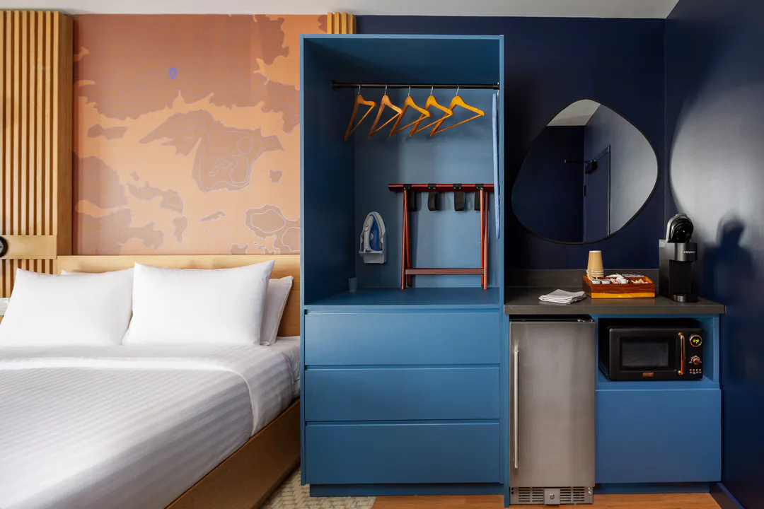 A modern hotel room with a bed, a large blue closet unit containing an iron and luggage rack, and a counter with a mini-fridge, microwave, and coffee maker, all set against a dark blue wall with an asymmetrical mirror.