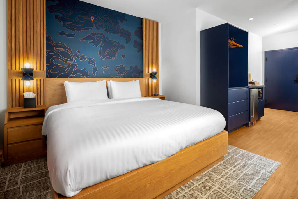 A bright, modern hotel guest room features a central bed with white linens against a headboard with a blue topographical map mural. To the right is a blue wardrobe unit with a small built-in beverage station, wood floors, and a textured area rug.