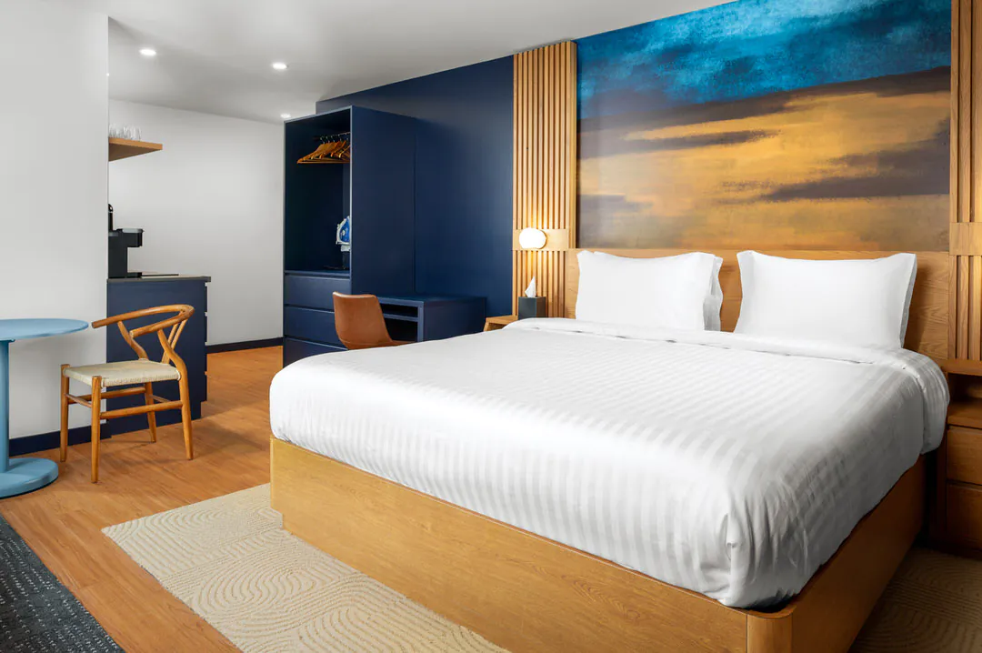 A modern hotel room with a large bed against a blue feature wall displaying landscape art. The room includes a wooden bed frame, a small dining table with a wooden chair, and a wardrobe closet with a coffee station in the corner.