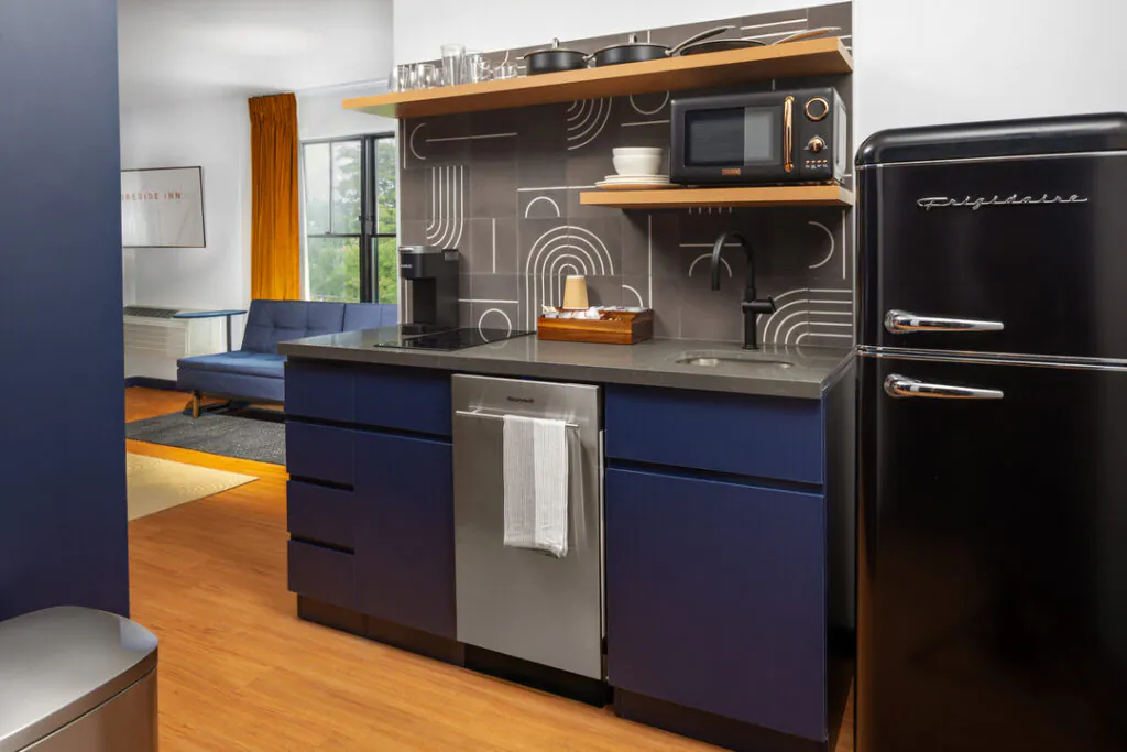A modern kitchenette with deep blue cabinets, a stainless steel dishwasher, and a black retro-style Frigidaire refrigerator. The area includes a sink, coffee maker, and a microwave on shelving against a decorative geometric tile backsplash.