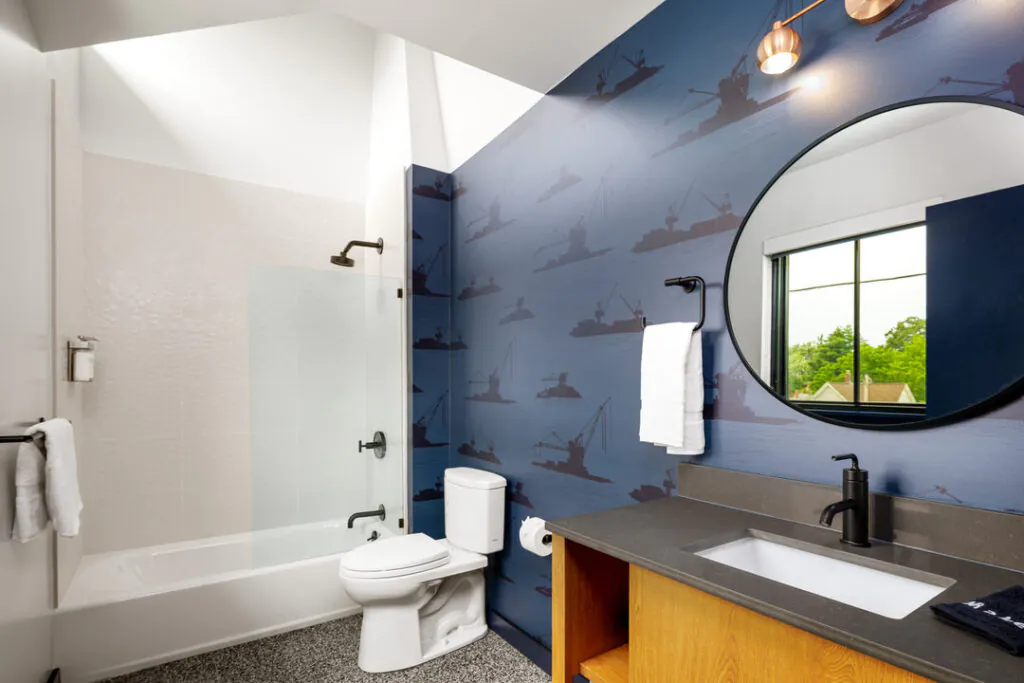 A modern bathroom featuring blue wallpaper with repeating crane and ship illustrations, a white toilet, a walk-in shower with a glass panel, and a wooden vanity topped with a dark gray counter, a round mirror, and black matte fixtures.