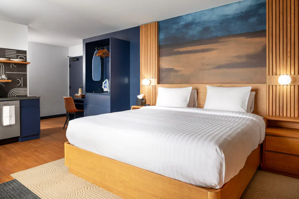 A modern hotel room featuring a large, neatly made bed with white linens, set against a dark blue and wood-paneled wall with warm accent lighting. To the left is a compact kitchenette, a small desk, and a built-in wardrobe with an ironing board.