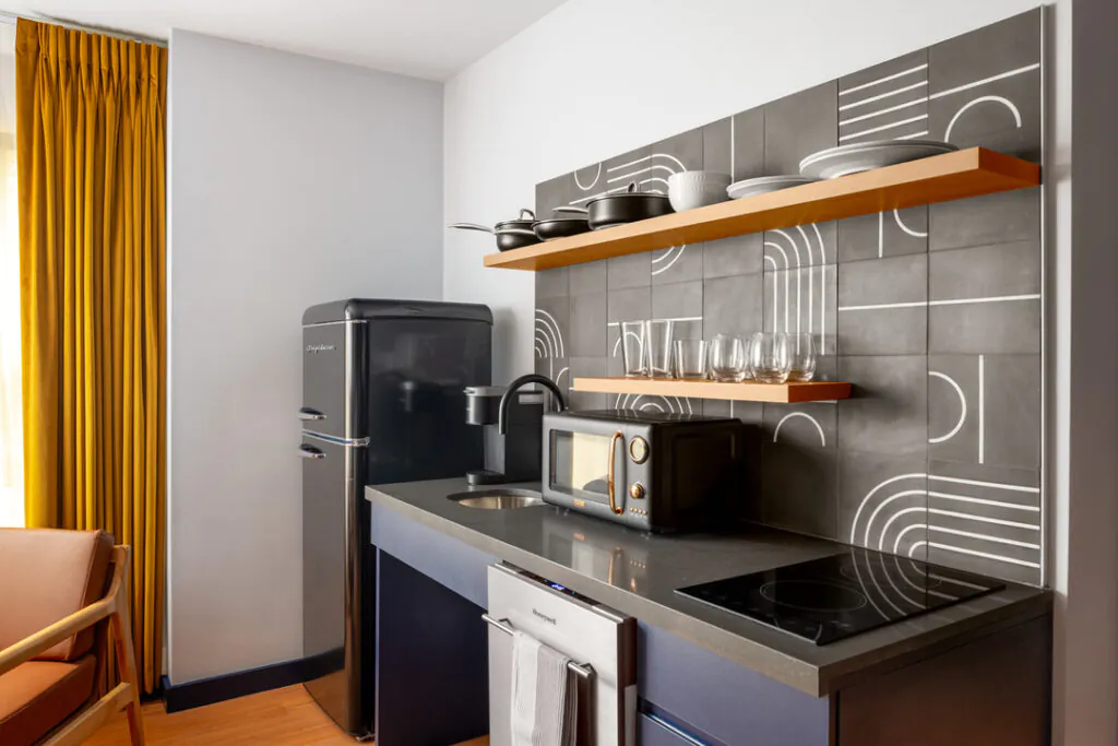 A modern kitchenette featuring a black retro refrigerator, grey countertops with a built-in microwave, sink, and induction cooktop. Floating wooden shelves display glassware and dishes against a stylish grey and white geometric tile backsplash.