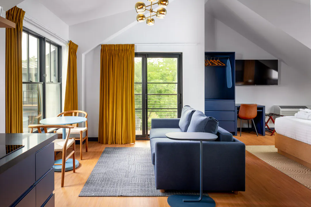 A modern hotel room featuring a blue sofa on a grey rug, a round dining table with two chairs by windows with mustard yellow curtains, a navy blue desk area with a brown chair, a gold modern ceiling light, and a glimpse of a wooden bed frame.