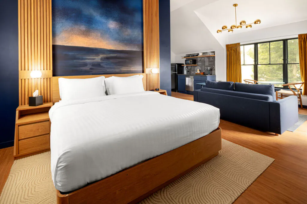 A modern hotel room featuring a plush king-sized bed with white bedding, framed by wood paneling and a large abstract painting. In the background, there is a blue sofa, a small kitchenette, and a window with yellow curtains letting in daylight.