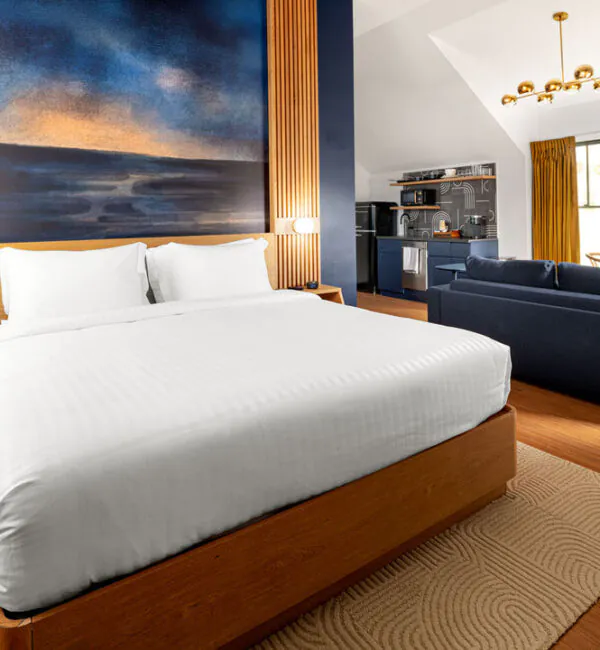 A modern hotel room featuring a plush king-sized bed with white bedding, framed by wood paneling and a large abstract painting. In the background, there is a blue sofa, a small kitchenette, and a window with yellow curtains letting in daylight.