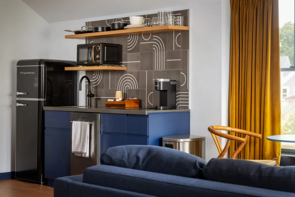 A modern kitchenette features a black retro refrigerator, blue cabinets, and a grey countertop. A decorative geometric-patterned backsplash sits behind a sink, microwave, and coffee station. To the side, a wooden chair sits near yellow curtains.