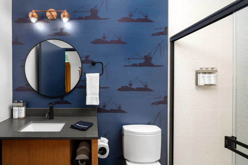 A modern bathroom interior featuring deep blue wallpaper with dark blue crane illustrations, a round mirror, a wooden vanity with a dark countertop, a white toilet, and a glass-enclosed shower stall with wall-mounted soap dispensers.